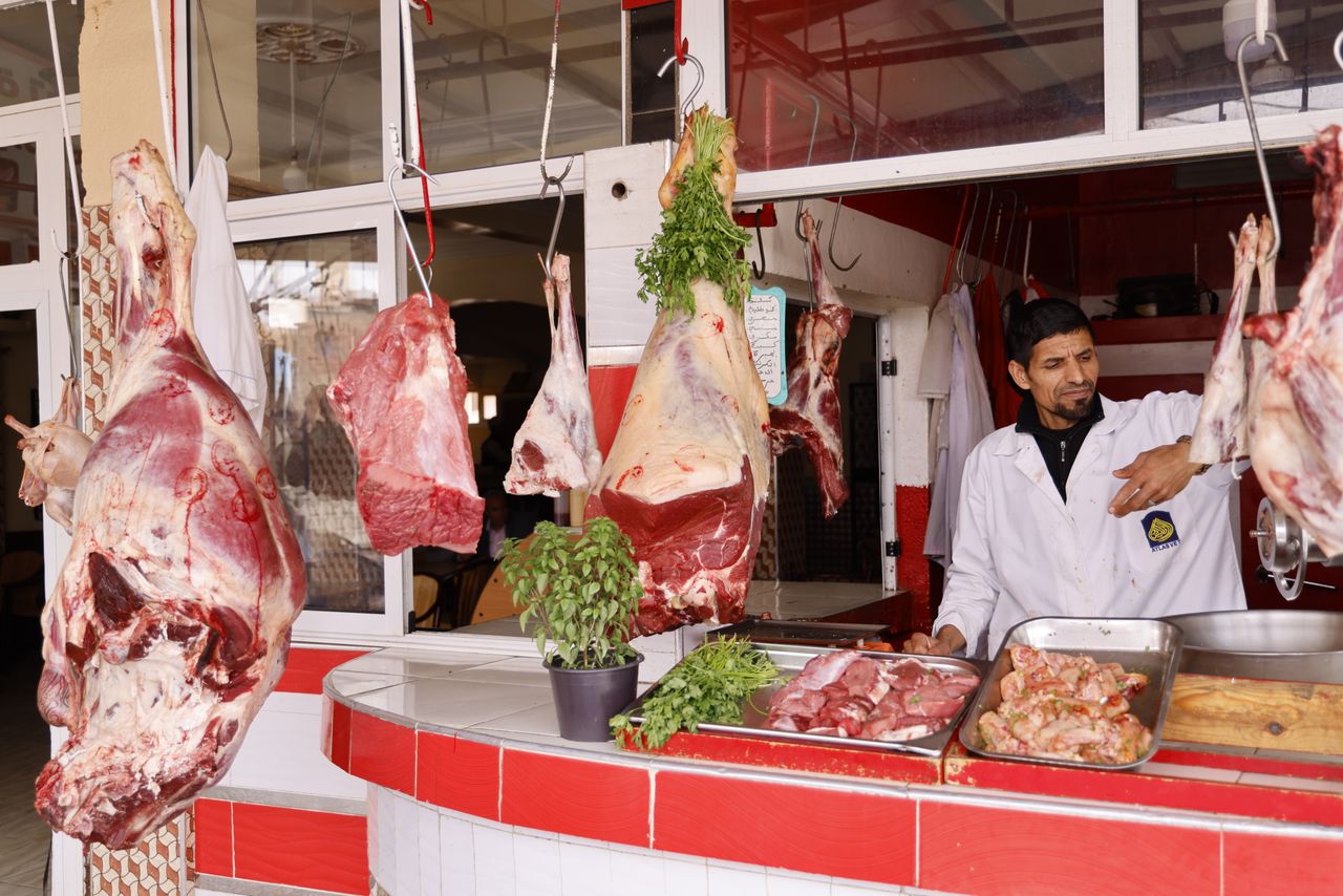 ours-journey A butcher working at an outdoor market stall with various cuts of beef hanging, showcasing fresh meat preparation.