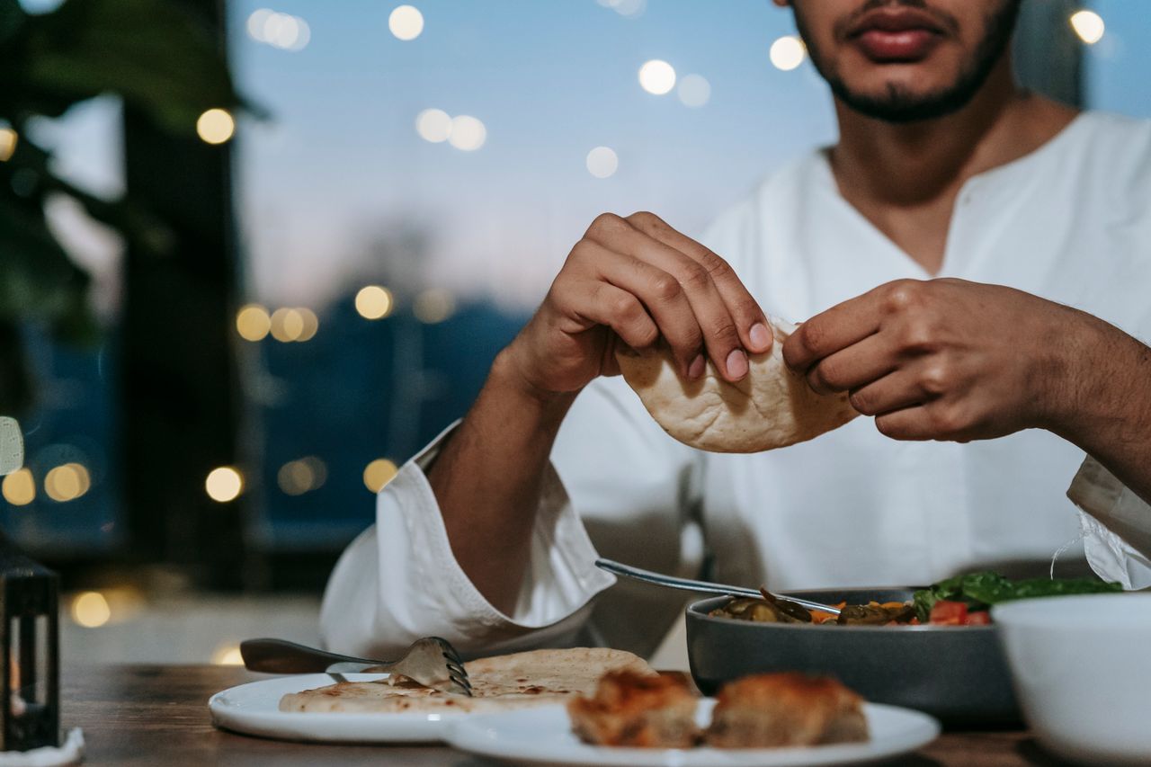services-01 Muslim man breaking pita bread at a dining table for dinner during iftar indoors.